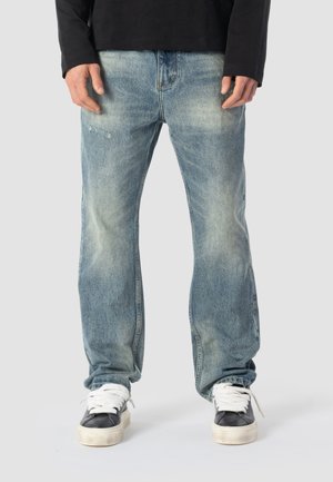 Mand iført løse, falmede blå jeans og sorte og hvide sneakers, står mod en ensfarvet grå baggrund.