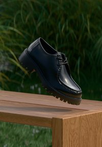 Chaussure noire en cuir à lacets avec semelle épaisse à rainures, flottant au-dessus d'un banc en bois en plein air avec un fond de feuillage vert.