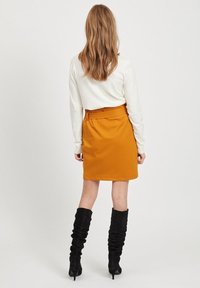 Jupe crayon orange avec une taille structurée et un détail ceinture, associée à un haut blanc à manches longues et des bottes noires à talons montantes jusqu'aux genoux.