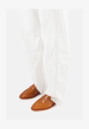 Brūni ādas loafers ar izšūtu dizainu, sudraba aksesuāriem un noapaļotu purngali, kombinēti ar baltām, brīvi pieguļošām biksēm.