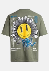 Camiseta verde oliva con un gráfico de un girasol sonriente con un centro amarillo, pétalos negros y texto azul goteante, además de acentos impresos.