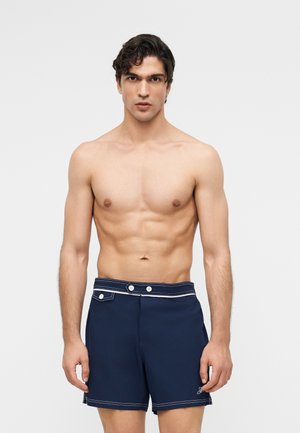 Oben ohne stehender Mann mit lockigem dunklem Haar, der marineblaue Badeshorts mit weißen Nähten und Knöpfen trägt, vor einem schlichten hellen Hintergrund.