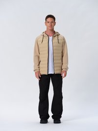 Beige gewatteerde jas met een capuchon, voorrits en trekkoorden, gecombineerd met zwarte broek, gedragen over een wit T-shirt en zwarte schoenen.