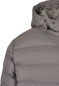 Grå vadderad jacka med axel- och huva samt justerbar dragsko och knäppning på vit bakgrund.