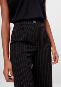 Svarta smalrandiga byxor med hög midja, enkel knappstängning och veckad framdesign. Tygets struktur verkar jämn och ställd.