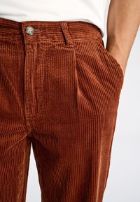 Pantalones de pana marrón con canalé vertical, con cierre frontal de botones y bolsillos laterales. Textura suave y ajuste relajado.