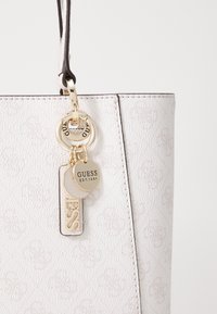 Sac à main texturé blanc avec quincaillerie dorée, présentant une étiquette porte-clés portant l'inscription "GUESS" et un motif logo en relief sur la surface.