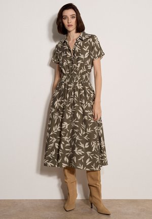 Donna che indossa un vestito olivastro a maniche corte con motivi di foglie bianche, vita stretta e stivali con tacco alti fino al ginocchio color marrone chiaro, in piedi contro un muro semplice.