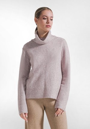 Kvinde iført en lyserød rullekravesweater og beige bukser, stående mod en ensfarvet hvid baggrund, kigger ned mod hendes højre side.
