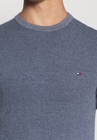 Sininen neulottu pyöreäkauluksinen villapaita, jossa on teksturoitu kangas ja pieni brodeerattu logo, jossa on punaisia, valkoisia ja sinisiä yksityiskohtia rinnassa.