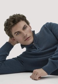 Marineblaues Zip-Pullover aus strukturiertem Strickstoff. Verfügt über einen hohen Kragen und umgeschlagene Ärmel. Das Model lehnt sich mit einer Hand hinter dem Kopf zurück.