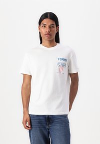 T-shirt en coton blanc avec imprimé graphique bleu comportant le texte "TOMMY" et un élément de design. Manches courtes, encolure ras du cou, coupe décontractée.