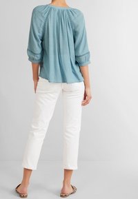 Blouse bleu clair, ample, à manches trois-quarts, tissu texturé, avec des détails froncés au dos, portée avec un pantalon blanc ajusté.