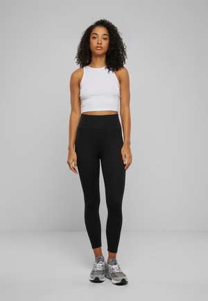 Sorte høye leggings og en hvit cropped tank top, begge i elastisk stoff; modellen har på seg grå joggesko. Enkelt design, tettsittende silhuett.