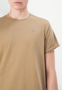 Kortermet t-skjorte med rund hals i bomull i tan-farge, med en subtil preget logo på brystet og opprullede ermer. Glatt tekstur.