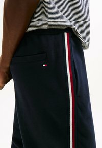 Mørkeblå sweatpants med sidestriber i rød, hvid og blå. Har en baglomme og et lille logo-patch i samme farveskema.
