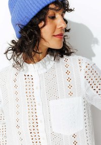 Blouse blanche en dentelle texturée avec des motifs à œillets, un col volanté et une poche poitrine. Un bonnet en côtes bleu complète le look.
