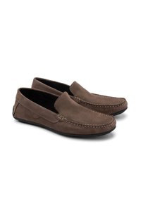 Mocasines de ante marrones con una textura suave, diseño sin cordones, costuras prominentes a lo largo de los bordes y una suela baja de goma negra.