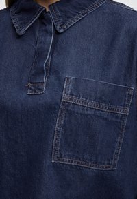 Mørk denimskjorte med spids krave, en halv knaplukning og en enkelt brystlomme med synlige syningsdetaljer.