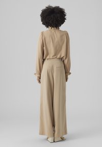 Blusa beige con scollatura arricciata e maniche lunghe, abbinata a pantaloni beige a vita alta e gamba larga. Tessuto dalla superficie liscia.