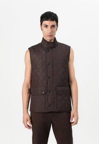 Καφέ επενδυμένο γιλέκο με ψηλό γιακά, αμάνικο σχέδιο, μπροστινό κλείσιμο με κουμπιά και δύο μεγάλες τσέπες με καπάκι. Απαλή υφή, μίνιμαλ λεπτομέρειες.