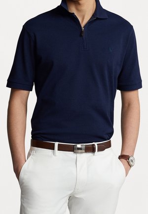 Man in een marineblauwe polo met korte mouwen en rits, witte broek, een bruine leren riem met metalen gesp en een bruine leren horloge met zilveren wijzerplaat.