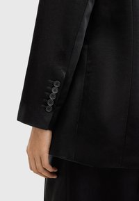 Manica di un blazer nero con cinque bottoni neri, dettagli di cucitura visibili e una finitura in tessuto testurizzato. Una mano che tiene il blazer è parzialmente visibile.