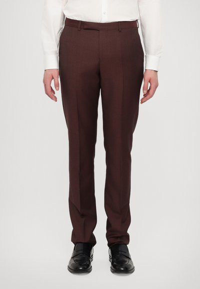 Pantalons marron ajustés avec un devant plat et des détails de pli, fabriqués en tissu lisse, associés à des chaussures foncées et une chemise blanche.