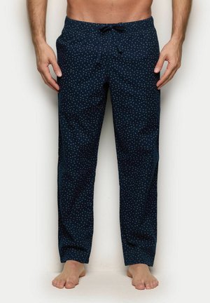 Uomo con pantaloni blu navy decorati con piccoli motivi di ancore bianche, in piedi a piedi nudi davanti a uno sfondo bianco semplice.