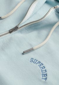 Sweat à capuche zippé bleu clair en tissu doux, avec des cordons de serrage blancs et un logo "Superdry" brodé dans un design incurvé. Détail de fermeture éclair en argent.