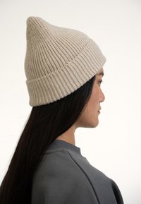 Béžová pletená čepice s špičatým vrcholem, s ribovanou strukturou a ohrnutým okrajem, kterou nosí osoba s dlouhými tmavými vlasy.