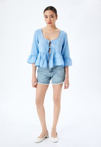 Lichtblauwe blouse met lange mouwen en een peplumzoom, vetersluiting aan de voorkant en geruffelde manchetten, gecombineerd met denim shorts en witte schoenen.