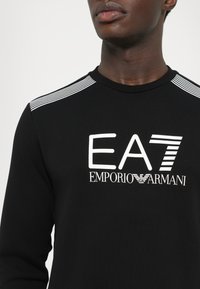 Black sweatshirt featuring a white EA7 logo, with horizontal white stripes across the shoulders and a ribbed crew neckline. Smooth texture.
