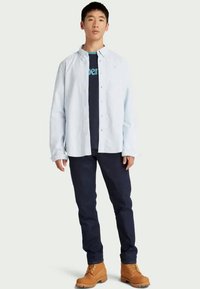 Camisa de manga larga azul claro sobre una camiseta oscura de color navy, combinada con pantalones oscuros y botas marrones con cordones. Enfocándose en las capas y un estilo casual.