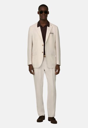 Mężczyzna ubrany w kremowy garnitur z brązową koszulą, ciemne okulary przeciwsłoneczne i brązowe buty, stojący na jednolitym jasnym tle.