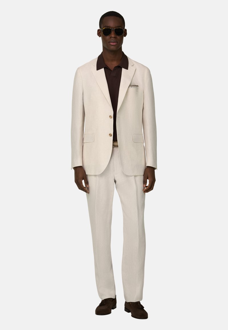 Homme portant un costume couleur crème avec une chemise marron, des lunettes de soleil foncées et des chaussures marron, debout devant un fond clair uni.