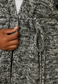 Stickad cardigan i svart och vitt med ett strukturerat mönster. Har ett knytbälte vid midjan och öppen frontdesign.