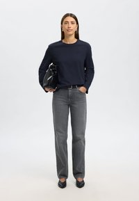 Mornarska košulja s dugim rukavima, sive izravne traperice, crne šiljate balerinke i crna torba s detaljem u čvoru. Model stoji ispred jednobojne pozadine.