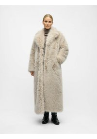Lang, lys beige faux pelsfrakke med en bred krave, forlommer og fuld længde. Modellen bærer en grå turtleneck og sorte støvler.