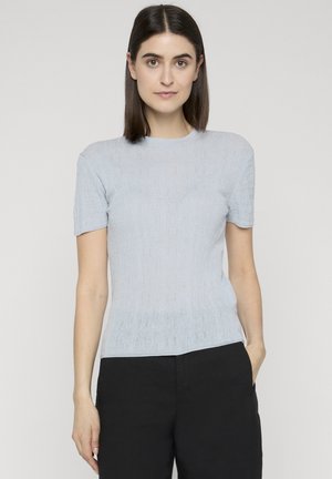 Frau mit glatten dunklen Haaren, die ein hellblaues Kurzarmshirt und schwarze Hosen trägt, steht vor einem schlichten hellen Hintergrund.