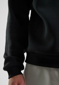 Schwarzer Sweatshirt mit gerippten Bündchen und Saum, der eine glatte, weiche Textur aufweist. Getragen über hellgrauen Hosen.