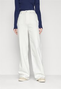 Jeans anchos blancos de cintura alta con bolsillos frontales, hechos de mezclilla. Combinados con una camiseta ajustada de manga larga en color azul marino y zapatillas beige.
