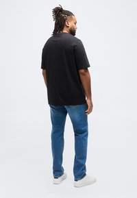 Άνδρας με κότσο dreadlocks που φοράει μαύρο μπλουζάκι, μπλε τζιν και λευκά αθλητικά, στέκεται με την πλάτη προς τα εμπρός σε ένα απλό ανοιχτό φόντο.