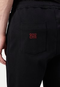 Zwarte joggers met een achterzak en rode geborduurde tekst "OPEN ERA." Glad materiaal met een elastische tailleband. Eenvoudig en functioneel ontwerp.