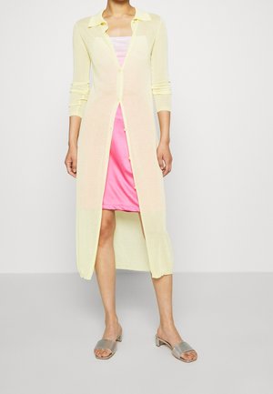 Lichtgele doorzichtige cardigan met een kraag, lange mouwen en knoopsluiting, gelaagd over een roze slipdress, aangevuld met zilveren slide sandalen.