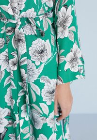 Blomstret kimono med en grøn baggrund, der har store hvide og sorte blomster. Lange ærmer, en bindebeltelukning og en glat tekstur.