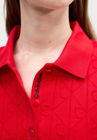 Gros plan sur un col de chemise rouge à motifs avec un texte noir "Calvin Klein" près des boutons, porté par une personne à la peau claire et aux cheveux bruns.