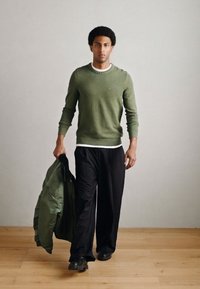 Žalias dryžuotas megztinis, juodos plačios kelnės ir žalia bombonešio striukė. Modelis laiko striukę, dėvi juodus batus, stovi ant medinių grindų.