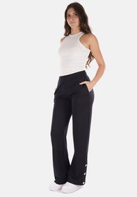 Pantaloni neri con vita elastica, tasche e dettagli con bottoni dorati alla caviglia, abbinati a una canotta bianca aderente e scarpe bianche.