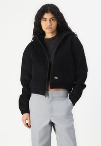 Giacca nera corta con chiusura a zip e polsini elasticizzati, caratterizzata da un piccolo logo cucito. Il modello indossa pantaloni grigi sotto.